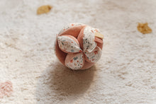Load image into Gallery viewer, A soft baby toy in the shape of a ball with floral patterns and pink ribbed sections, featuring a label and a small rattle.