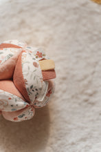Load image into Gallery viewer, A soft baby toy in the shape of a ball with floral patterns and pink ribbed sections, featuring a label and a small rattle.