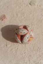 Load image into Gallery viewer, A soft baby toy in the shape of a ball with floral patterns and pink ribbed sections, featuring a label and a small rattle.
