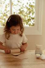 Load image into Gallery viewer, Child playing with a rolling pin on a wooden table near a window with greenery outside.
